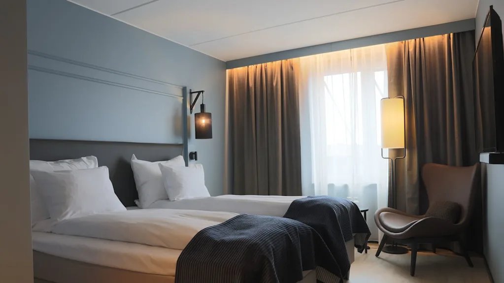 En pænt redt seng i et moderne hotelværelse med blødt lys, gardiner trukket for, og en hyggelig stol ved vinduet.