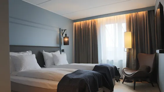A neatly made bed in a modern hotel room with soft lighting, curtains drawn, and a cozy chair by the window.