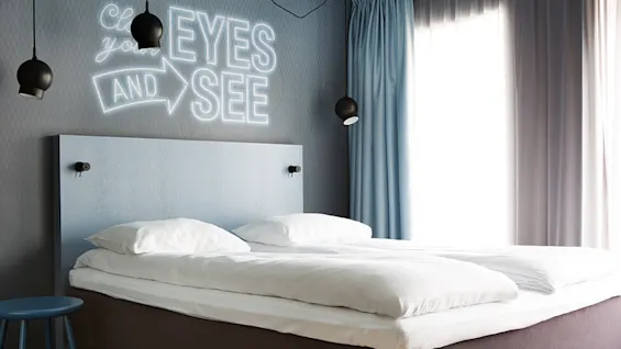En pent sengt med hvite sengetøy står i et moderne rom med blå detaljer. På veggen står det i neontekst: "Close your eyes and see". To svarte pendellamper henger over.