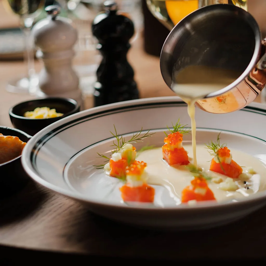 En person häller sås från en kopparpanna över en rätt med garnerade laxkuber på ett bord med salt-, pepparkar, bröd och glas.