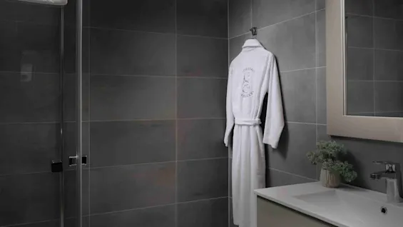 En hvid badekåbe hænger på en grå flisevæg ved siden af en vask med en plante, under skarpt lys i et moderne badeværelse.
