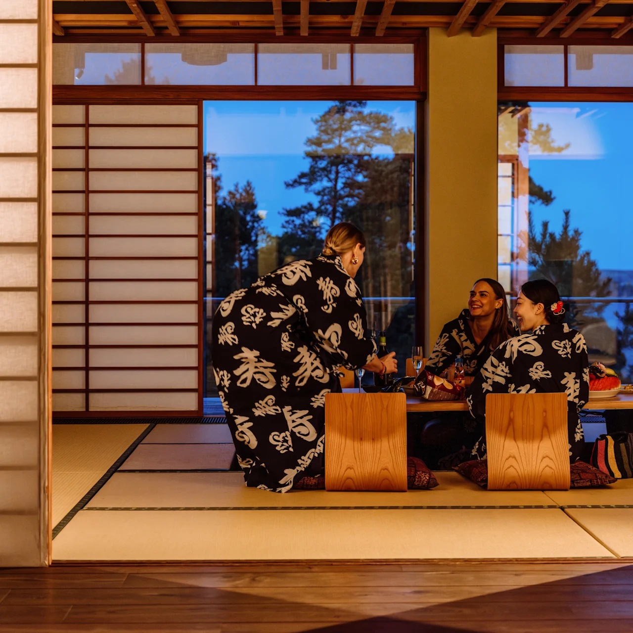 Tre personer i kimonos sitter og samtaler rundt et lavt bord. Rommet har shoji-skjermer og utsikt over trær gjennom store vinduer.