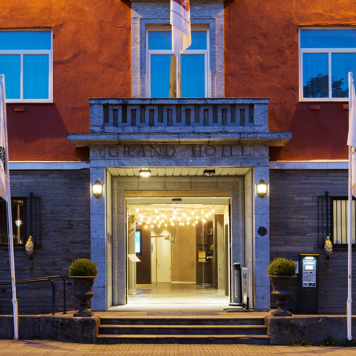 Grand Hotel-inngang med opplyste pærer hengende over, omgitt av en rød og grå utside. Steg leder opp til døren. Flagg står på hver side.