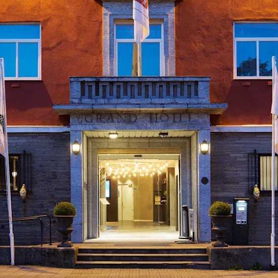 Grand Hotel-inngang med opplyste pærer hengende over, omgitt av en rød og grå utside. Steg leder opp til døren. Flagg står på hver side.