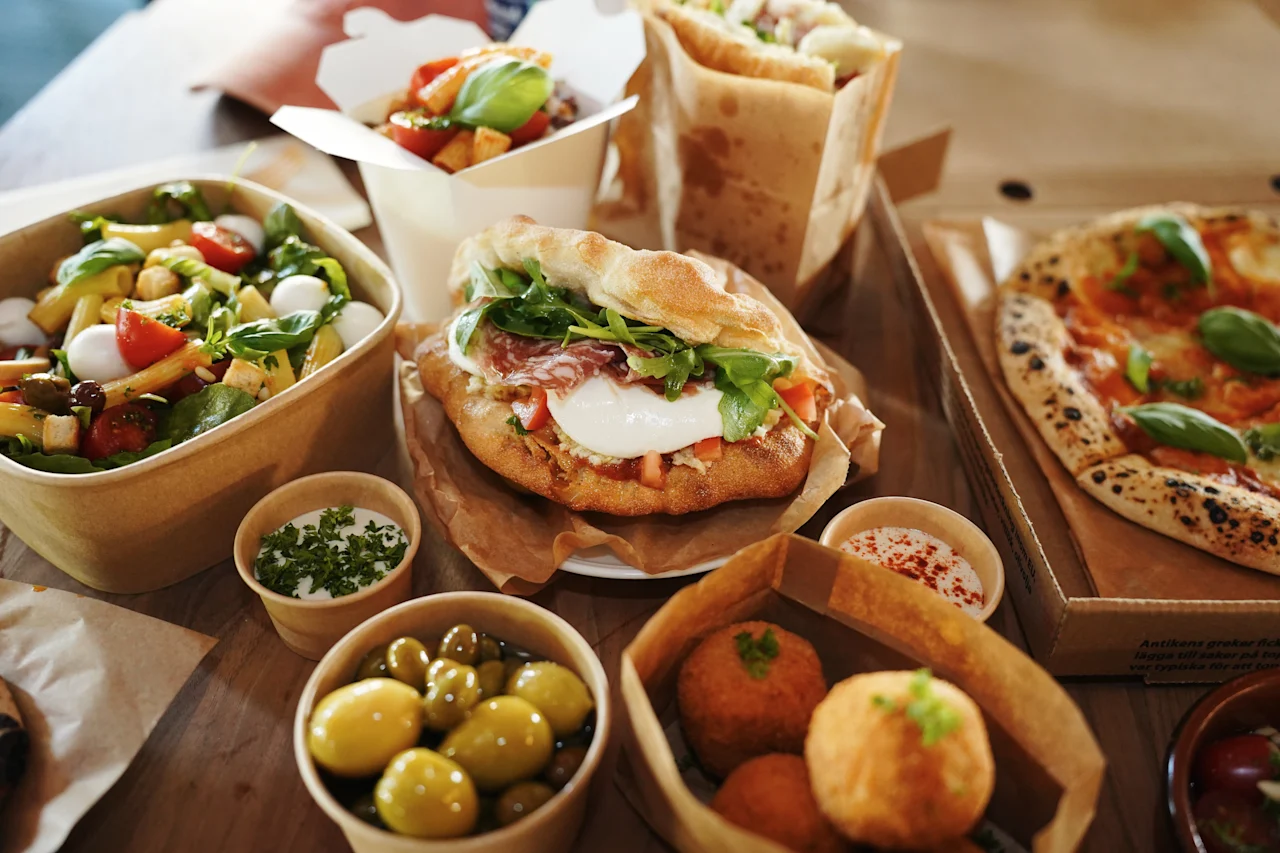Erilaisia italialaisia ruokia, kuten pizzaa, salaattia, voileipiä ja oliiveja, on asetettu puupöydälle, mikä viittaa rentoon ruokailu- tai takeout-tilanteeseen.