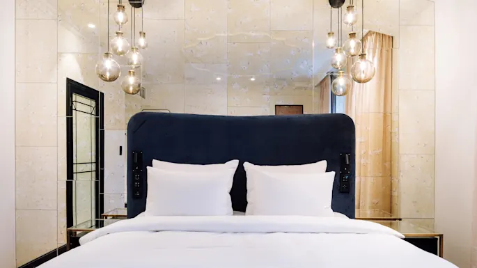 A bed with white linens and a dark headboard is centered under hanging globe lights in a modern, mirrored room with beige walls and a curtain.