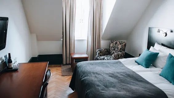 Seng med turkis puder står mod en væg, overfor en mønstret lænestol og vindue dækket af beige gardiner, i et hyggeligt værelse med TV og træskrivebord.