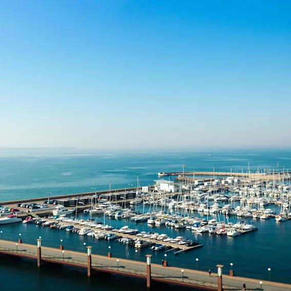 A marina filled with docked boats lies along a pier extending into a calm, expansive sea under a clear blue sky.