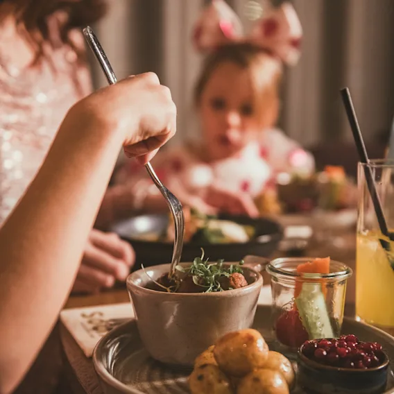 En mor som spiser kjøttkaker, poteter og tyttebær ved et restaurantbord med sitt lille barn i bakgrunnen.