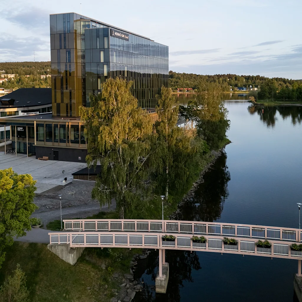 En moderne glasbygning reflekterer lyset, beliggende ved en rolig flod med omkringliggende træer og en lille gangbro. Tekst: "SBAB Gävle".