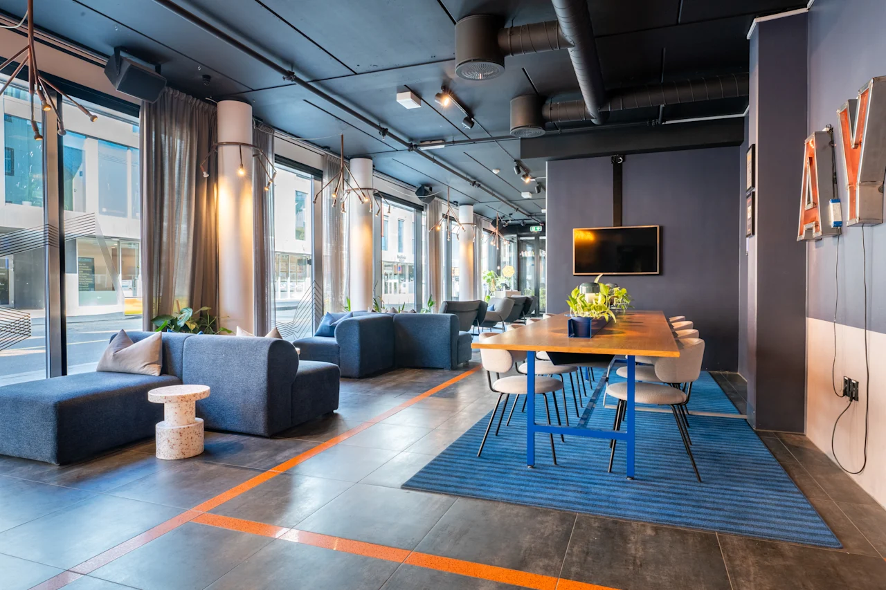 Et moderne loungeområde med et langt trebord og blå stoler plassert på et stripet blått teppe. Store vinduer langs veggen slipper inn naturlig lys rundt sofaer og inneplanter.