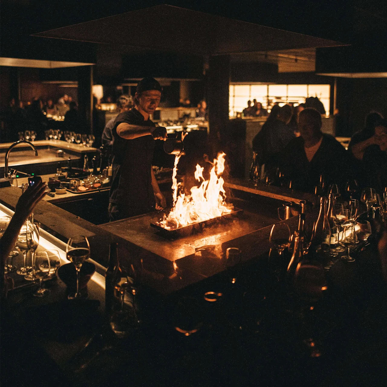 En kokk heller væske på en grill, noe som forårsaker flammer, i en dempet belyst restaurant.