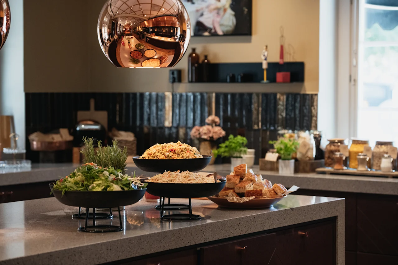 En buffet med friske salater, varme retter og brød på en disk, belyst av en reflekterende lampe.