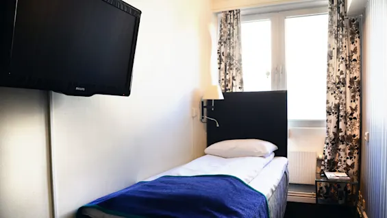 En enkeltseng med et blått teppe i et lite rom, med en TV montert på veggen og åpne gardiner som slipper inn dagslys.