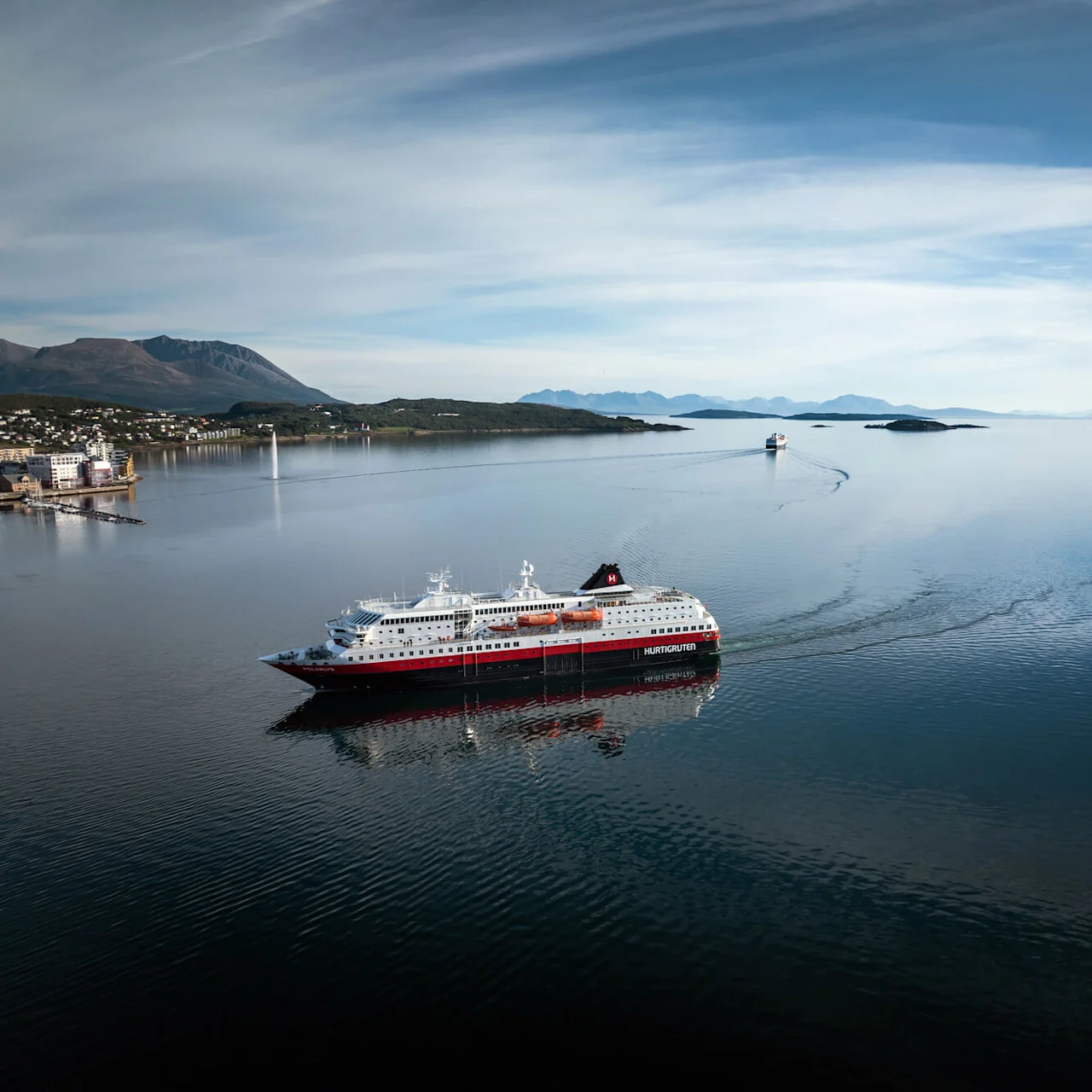 Krydstogtskib mærket "Hurtigruten" sejler gennem roligt vand og efterlader et vågen. Bjerge og en lille kystby er synlige i baggrunden under en delvis overskyet himmel.