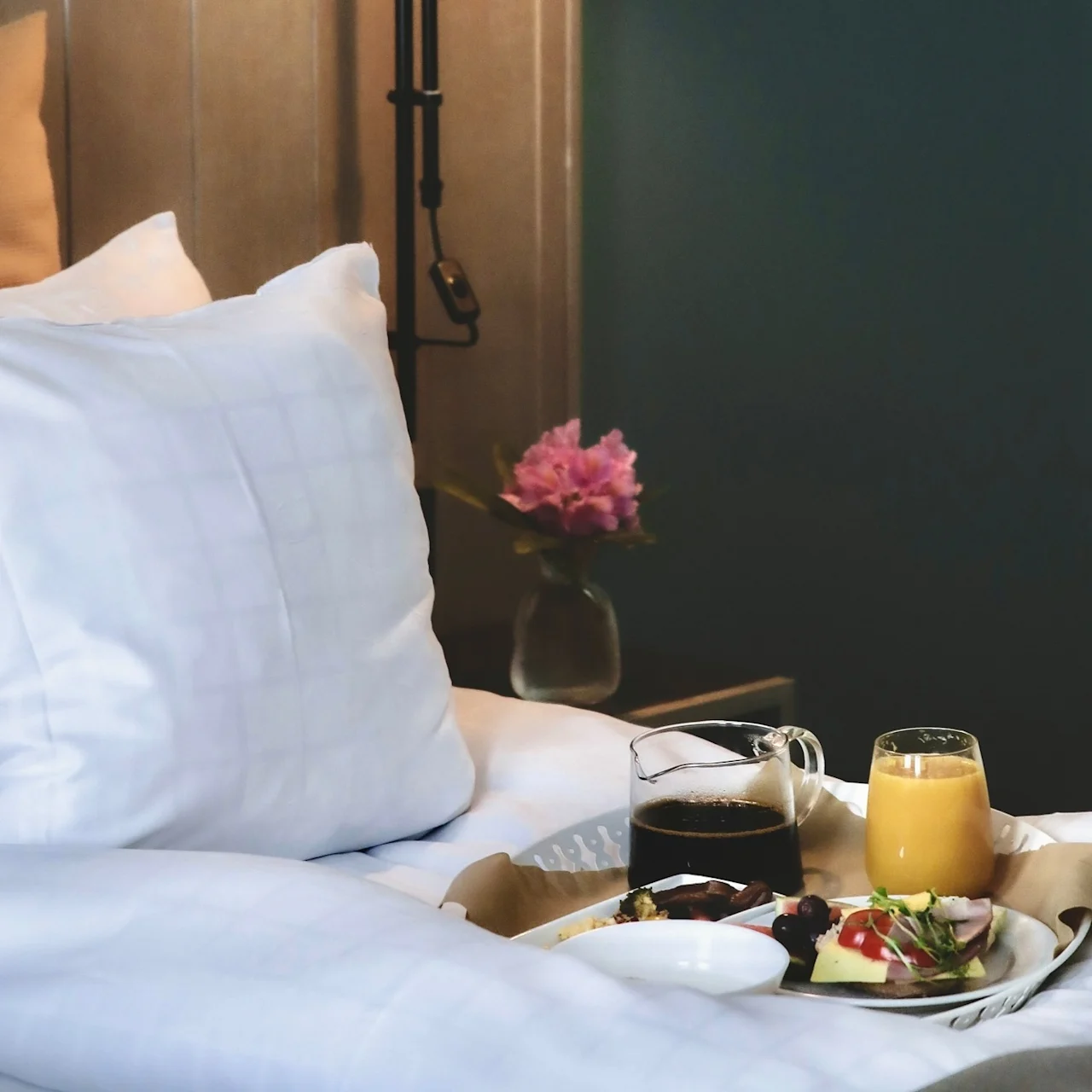 Breakfast tray rests on a bed with white pillows, holding coffee, juice, and a plate of food. A vase with pink flowers sits on a nearby table.