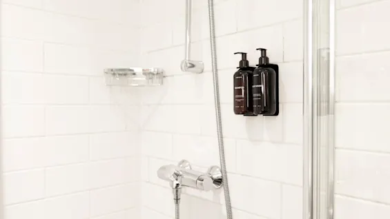 Shower area with white tiled walls, featuring shampoo and body wash dispensers. Home Hotel Grand Bodø.