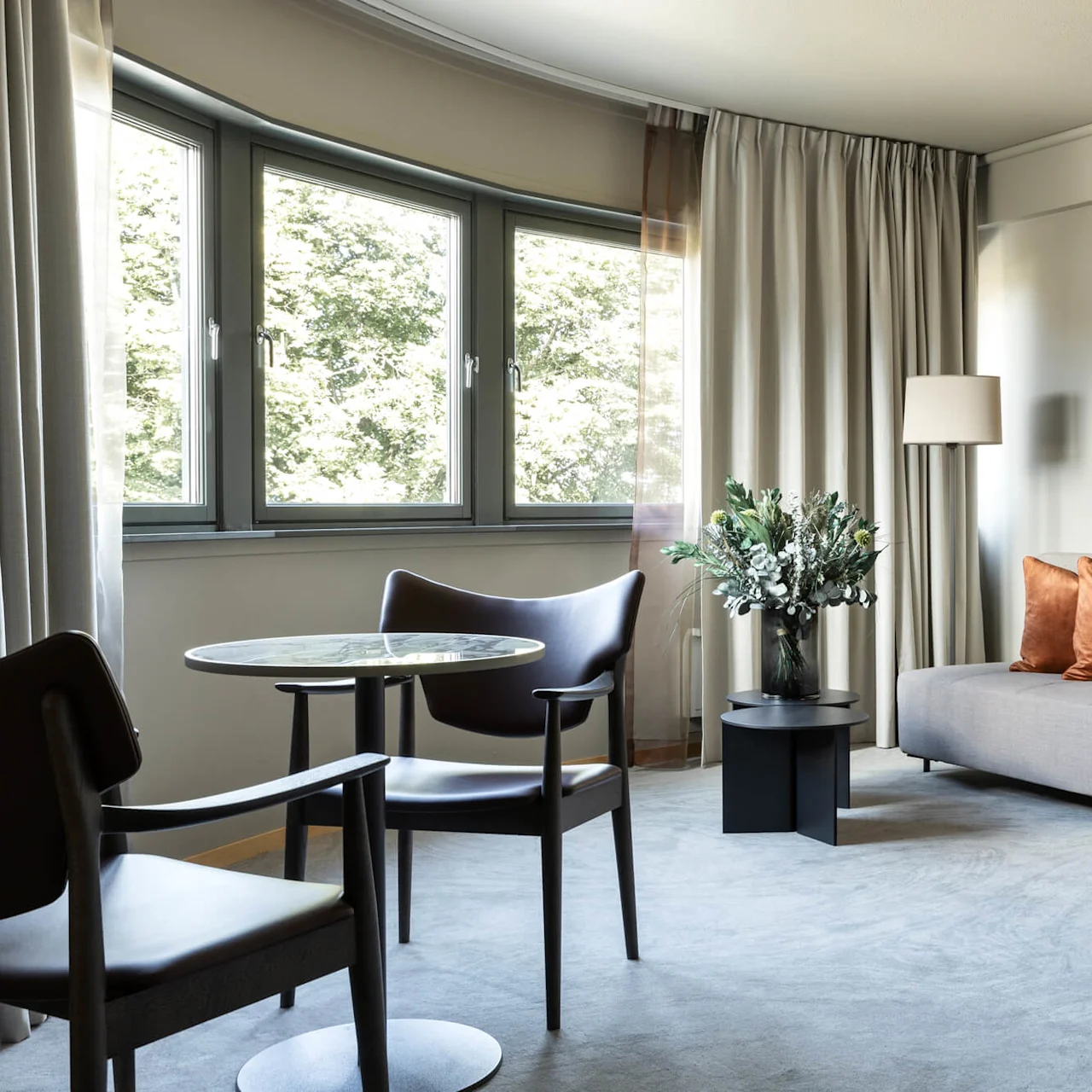 A gray sofa with orange pillows stands next to a tall lamp and a small table with flowers. Two black chairs are around a round table near large windows.