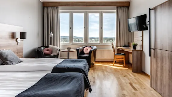 Ett prydligt hotellrum med två sängar med grå sängkläder, två fåtöljer vid ett fönster, en väggmonterad TV och ett skrivbord med en stol med utsikt över en naturskön vy.