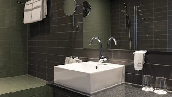 A white sink with chrome faucets situated in a modern bathroom; glass cups and a soap dispenser labeled "LUX" are nearby on a dark tiled wall.
