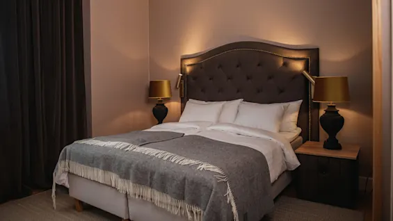 A neatly made bed with a tufted headboard, flanked by two dark lamps, rests in a warmly lit bedroom with brown walls and heavy curtains.