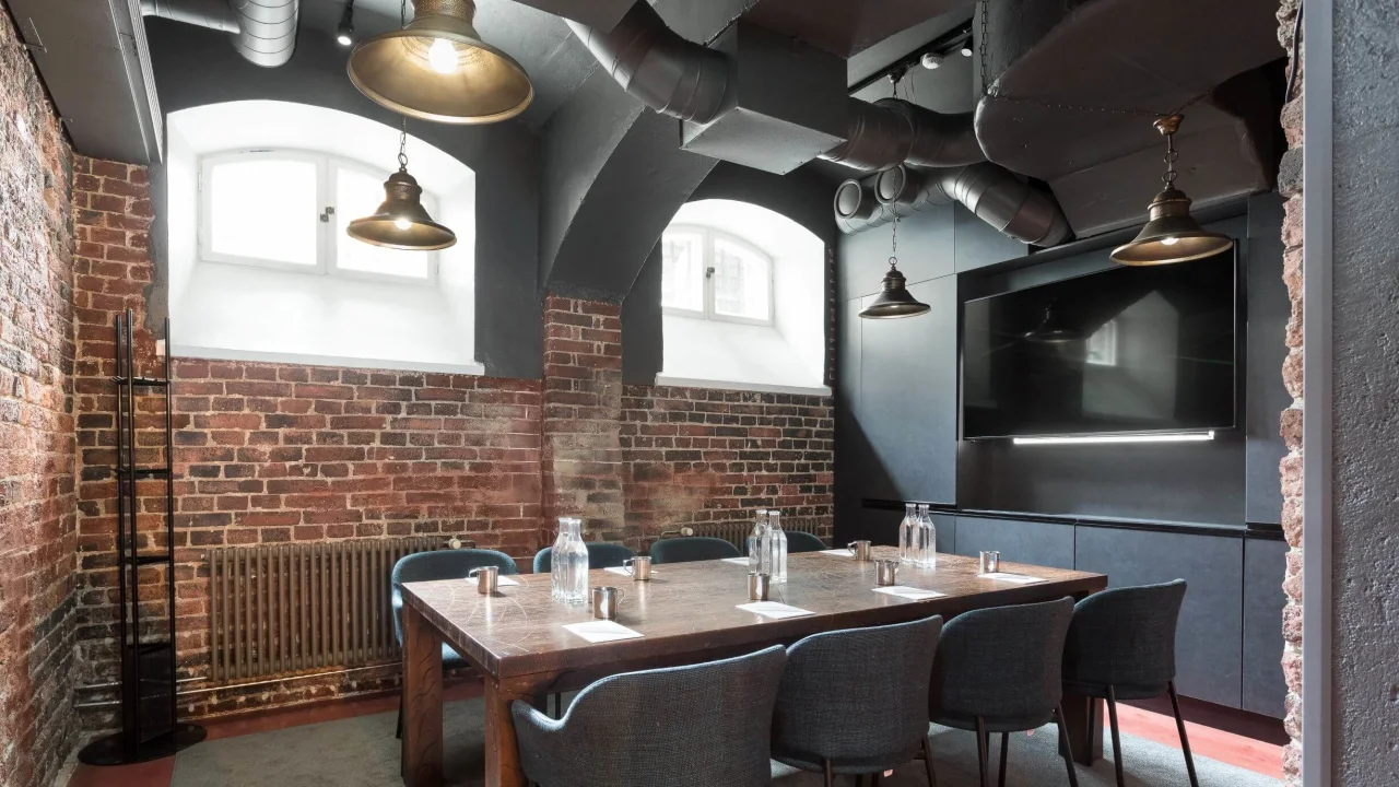 A dining area with a large table and chairs, under pendant lights, in a room with exposed brick walls.