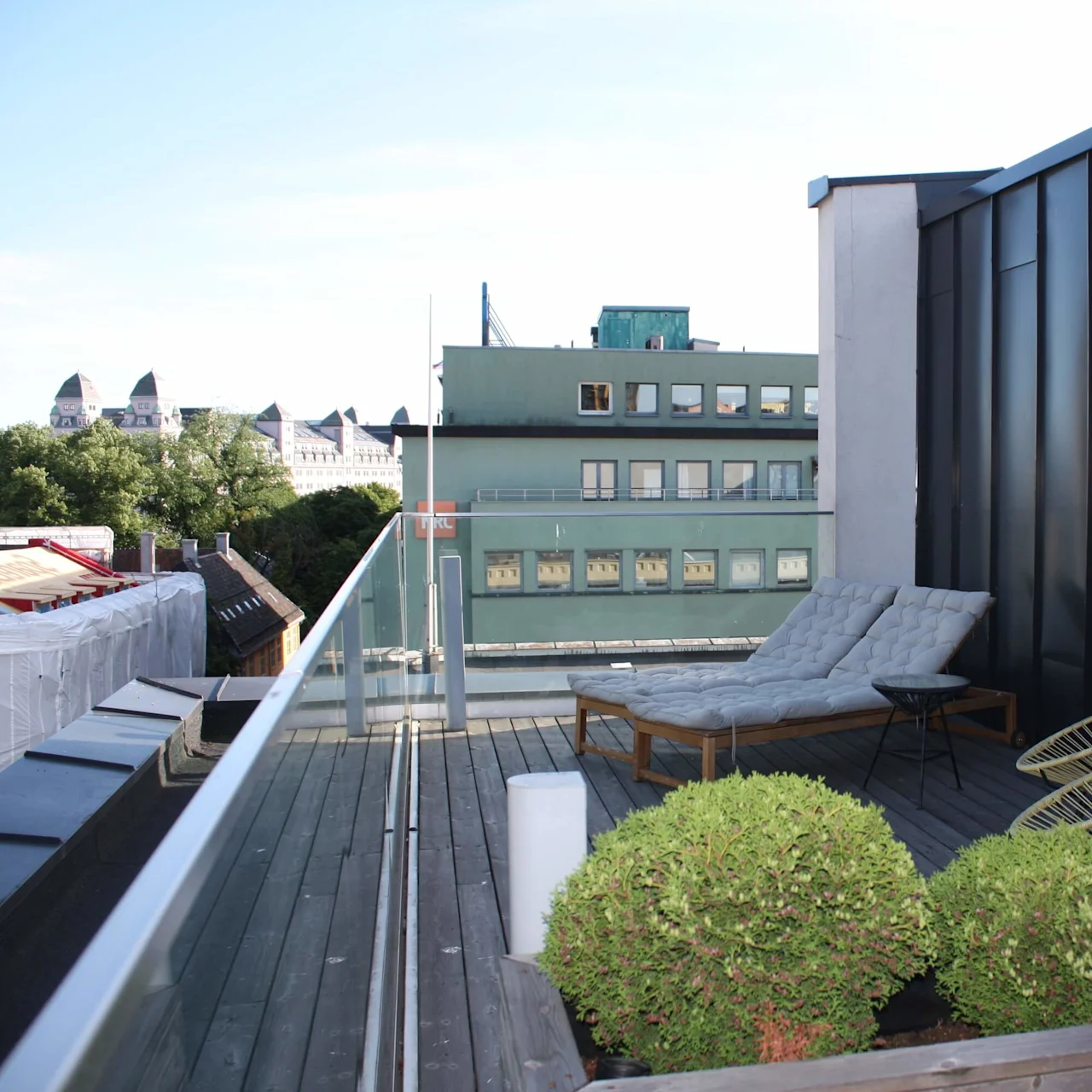 Rooftop terrace with two cushioned lounge chairs facing a glass railing; surrounded by bushes. Buildings and trees are visible in the background under a clear sky.