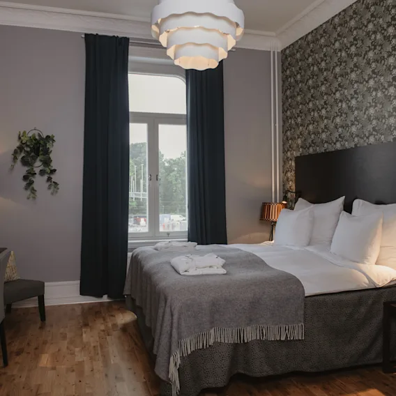 Ett bekvämt hotellrum har en prydligt bäddad säng med vita sängkläder mot en mönstrad vägg. Ett skrivbord och stol står bredvid under mjuk belysning. Ett fönster erbjuder utsikt utanför.