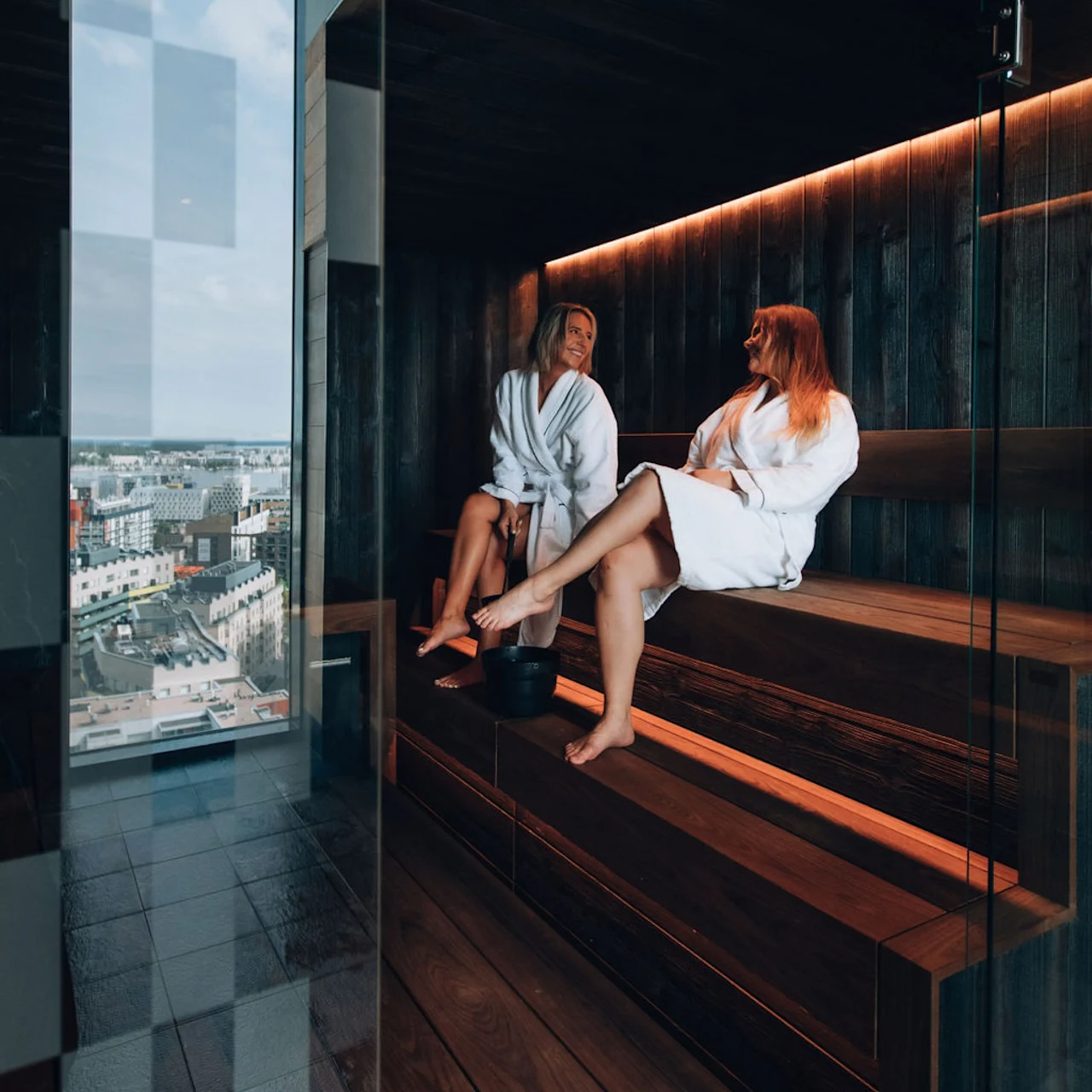 Kaksi kylpytakkiin pukeutunutta henkilöä rentoutuu ja keskustelee modernissa saunassa panoraamanäkymällä kaupunkiin.
