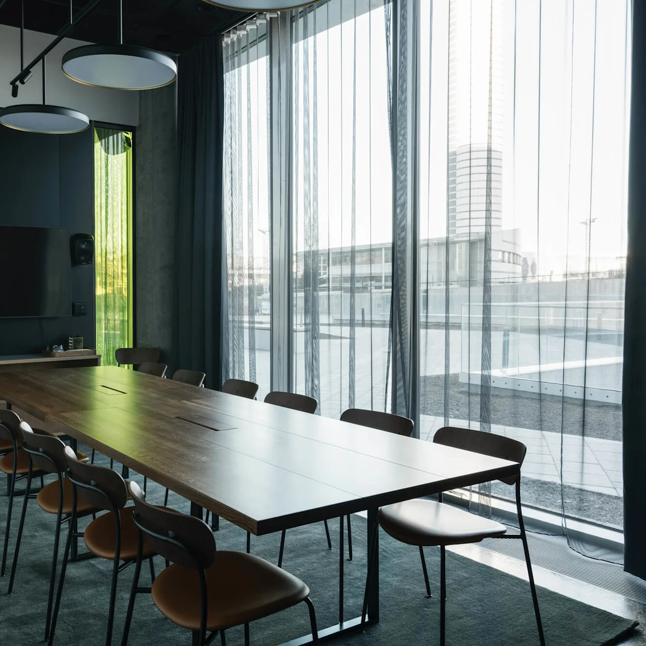 A large wooden conference table surrounded by chairs sits under modern ceiling lights in a room with floor-to-ceiling windows and sheer curtains, creating a bright and spacious setting.