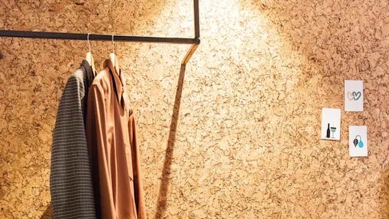 A beige chair sits on a dark carpet beside a wooden shelf. A coat and shirt hang on a rail above, with shoes below. Corkboard walls display small artworks.