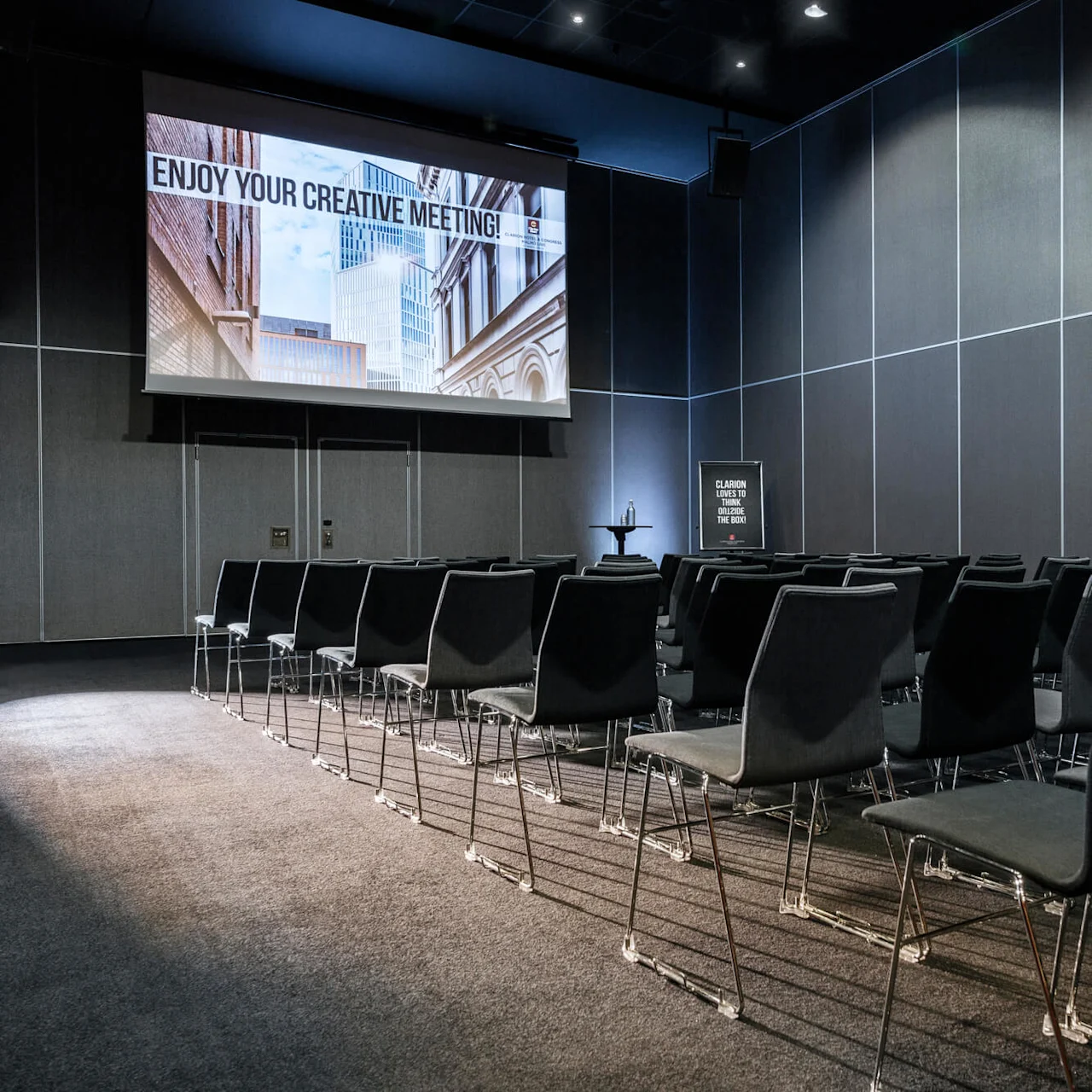 Rader med tomme stoler rettet mot en stor projeksjonsskjerm med teksten "ENJOY YOUR CREATIVE MEETING!" i et moderne konferanserom med grå vegger. Et skilt på siden leser "CLARION HOTELS THINK OUTSIDE THE BOX".
