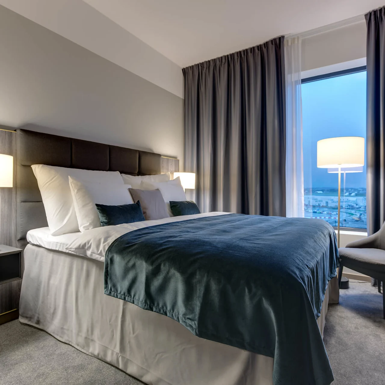 Seng med turkis sengetøy og hvite puter står mellom to tente lamper i et moderne hotellrom, ved siden av et stort vindu med lukkede gardiner og en lenestol.