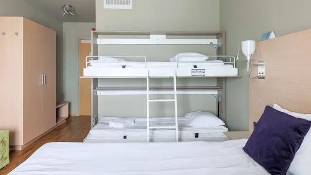 Våningssäng med vita sängkläder hopfälld mot en vägg. Stege leder till överslafen i ett minimalistiskt sovrum. Garderob och ljusa väggar kompletterar hotellrummets miljö.