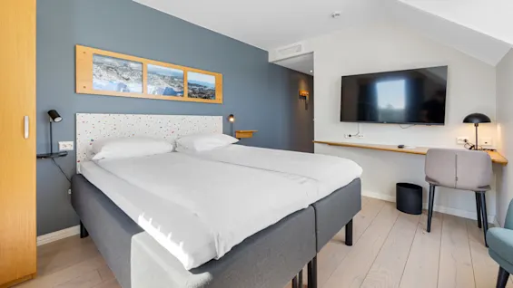 A neatly made double bed in a modern hotel room, flanked by artwork, a desk, and a flat-screen TV.