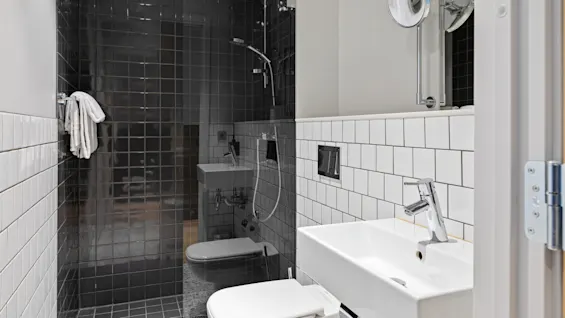 Et bad med en hvid håndvask og toilett, et spejl og et bruseområde med mørke fliser. Et håndklæde hænger på en stang. Væggene har kvadratiske fliser.