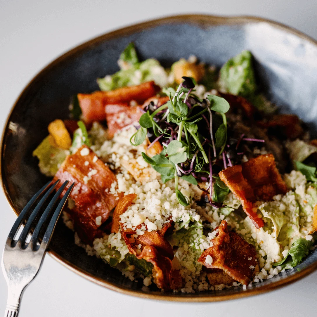 A salad with crispy bacon, croutons, and fresh greens sits appetizingly in a ceramic bowl on a white table, accompanied by a fork resting on the bowl's edge.