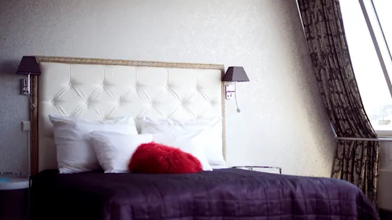 A plush, tufted headboard anchors a neatly made bed with crisp pillows and a red accent cushion, flanked by wall lamps, with a patterned curtain beside a bright window.