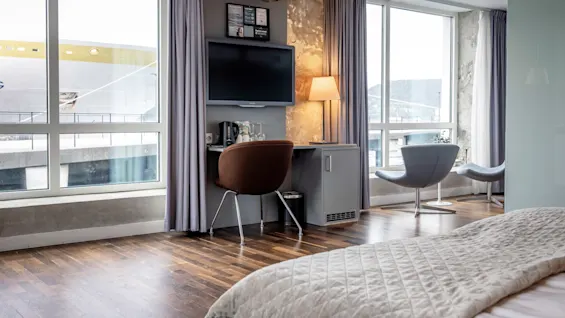 Hotellrum med säng, TV, lampa på skrivbord och stolar. Stora fönster visar utsikt över en förtöjd båt. Väggarna är grå och golvet i trä.
