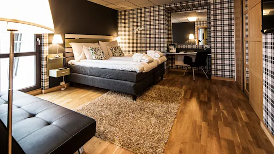 A neatly arranged hotel bedroom with a large bed, plaid walls, wooden floor, and a desk area.