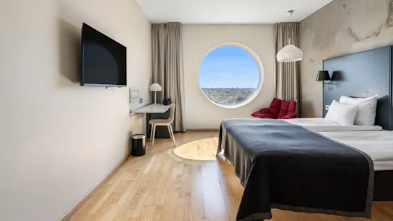 Moderne hotelværelse med et rundt vindue, der tilbyder byudsigt, en komfortabel seng, skrivebord og TV.