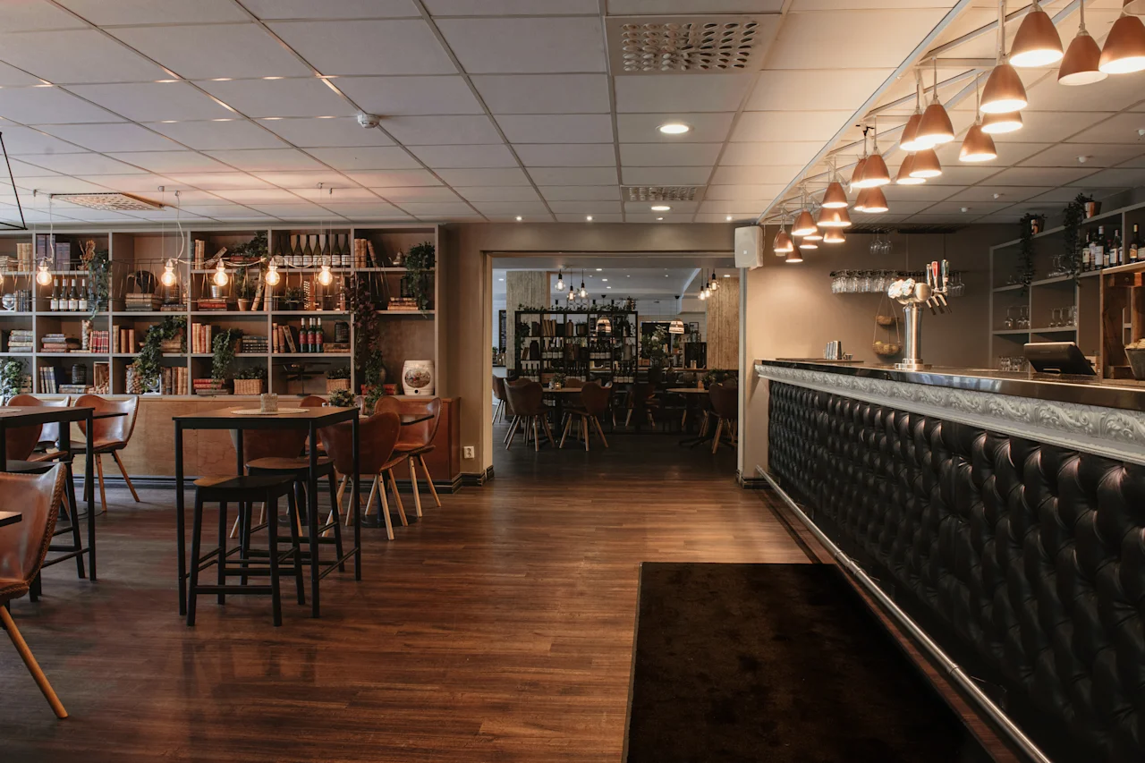 A cozy restaurant and bar area with warm lighting, wooden floors, a long bar, and bookshelves filled with books and plants.