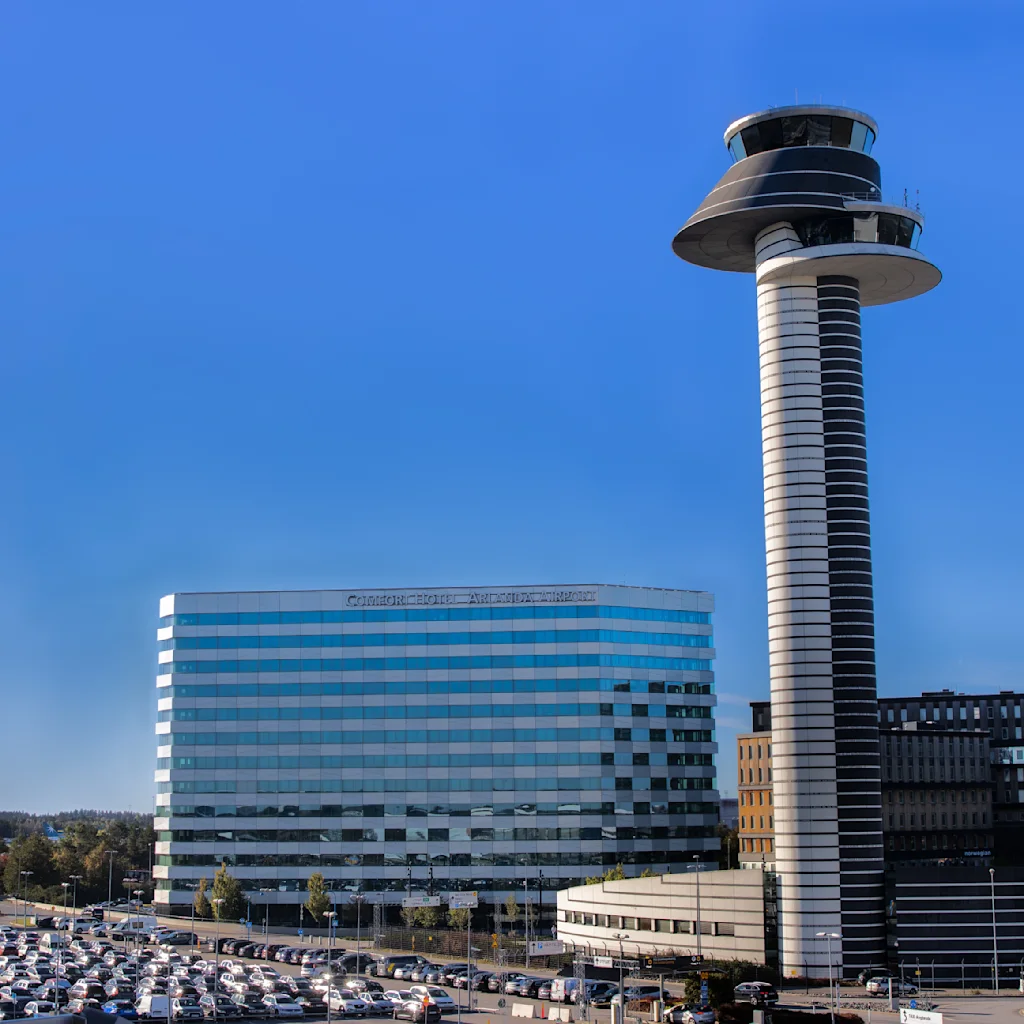 A modern hotel near the Arlanda Airport, highlighted by its sleek architecture, stands beside a distinctive control tower under a clear blue sky. Numerous parked cars fill the lot below.