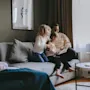 A family sits on a gray sofa, a parent reading a book to two children in a cozy living room with framed art and curtains.