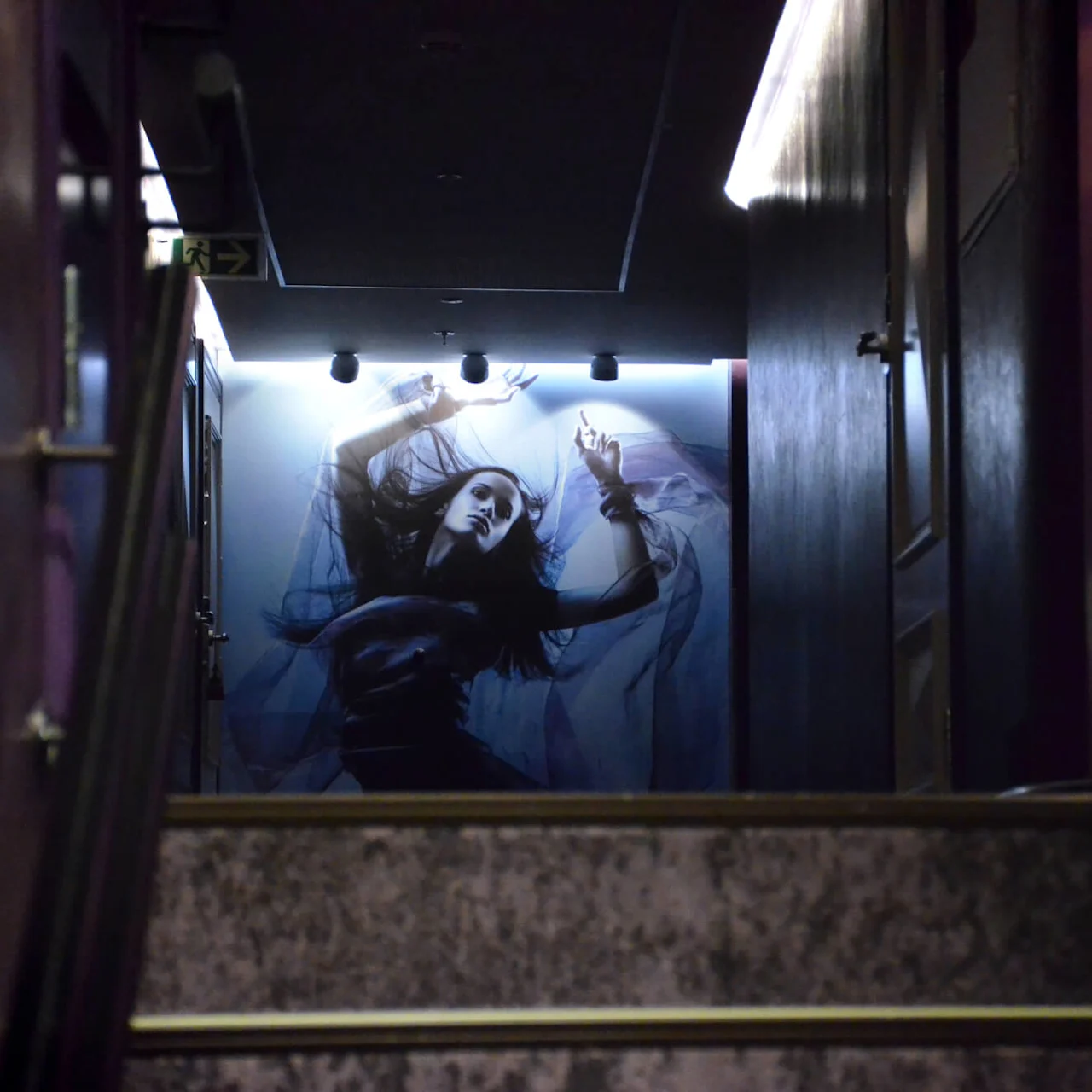 A large poster of a person in a dynamic pose adorns the wall at the top of dimly lit stairs, surrounded by dark walls and ceiling lights.