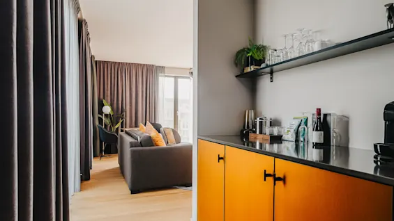 Et moderne opholdsområde med en grå sofa med orange puder, ved siden af høje vinduer og gardiner. Ved siden af et tekøkken med en hylde med glas og drikkevarer, fremhævet af et orange skab.