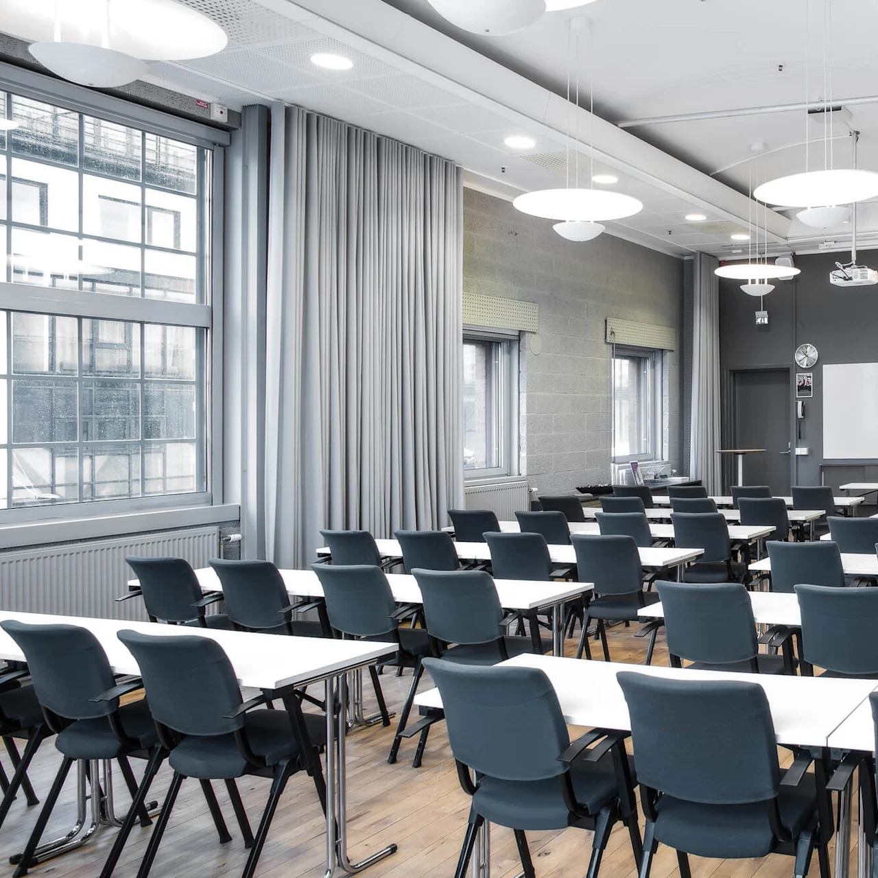 Classroom with rows of empty chairs and desks, facing a whiteboard. Large windows on the left, gray walls, and ceiling lights create a modern, well-lit environment.