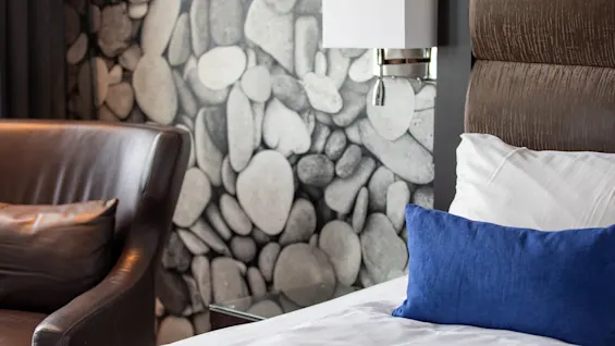 En seng med hvite og blå puter står ved siden av en brun skinnlenestol. Veggen bak har et steinmønster og en moderne vegglampe.