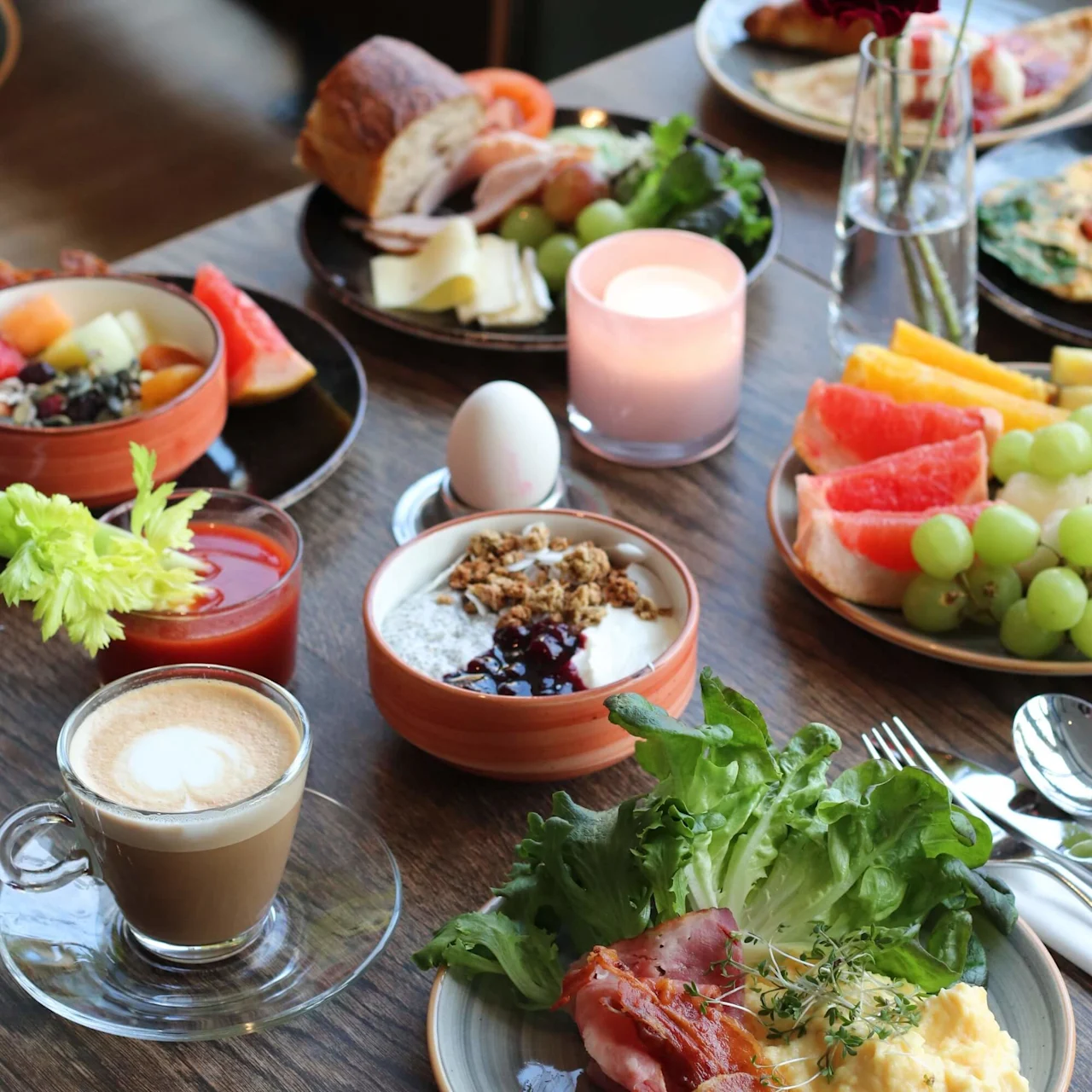 En frukost med äggröra, bacon, sallad, yoghurt med granola, färska frukter, en cappuccino och ett tänt ljus på ett träbord, skapar en varm och inbjudande atmosfär.