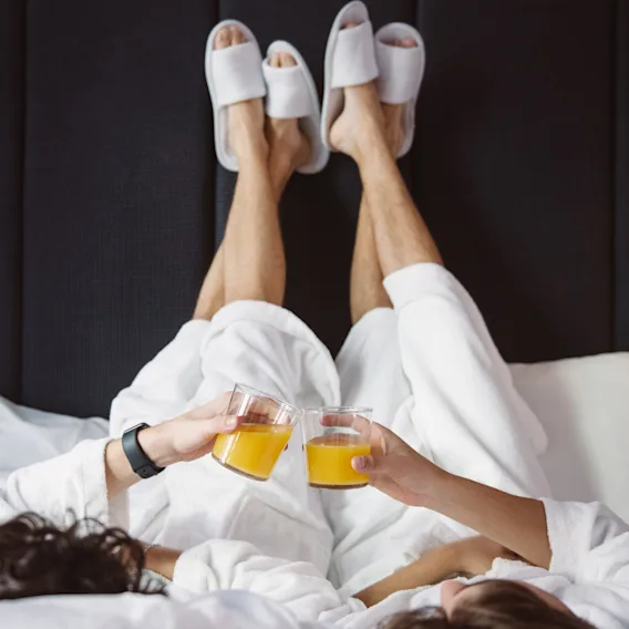 Två personer i badrockar och tofflor kopplar av i sängen och skålar med apelsinjuice. En mysig och glad stund av avkoppling.